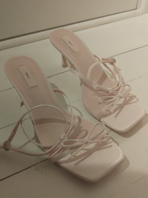 Pumps från Nelly - Säljer ett par ljusrosa pumps från Nelly med tunna remmar och söta rosetter framtill. Skorna har fyrkantig tå och smal klack, samt justerbar rem runt ankeln. Helt oanvända!