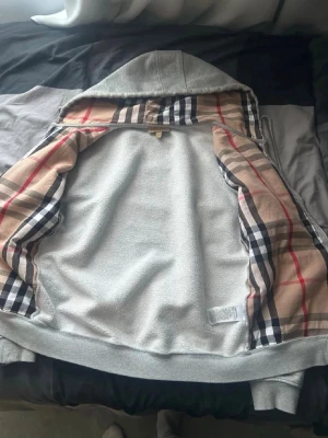 Grå hoodie med Burberry-mönster - Säljer en grå hoodie från Burberry med dragkedja och klassiskt rutigt foder i huvan och längs insidan. Hoodien har en mjuk bomullskänsla och ribbade muddar vid ärmar och nederkant. Perfekt för dig som gillar stilrena plagg med en twist.