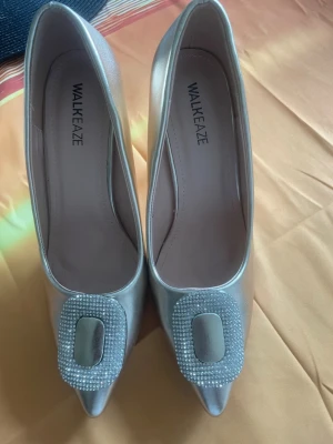 Silvriga pumps från WALKEAZE  - Snygga silvriga pumps från WALKEAZE med spetsig tå och dekorativt spänne med glittrande stenar framtill. Skorna har en elegant form och är tillverkade i ett skimrande material som ger en lyxig känsla.