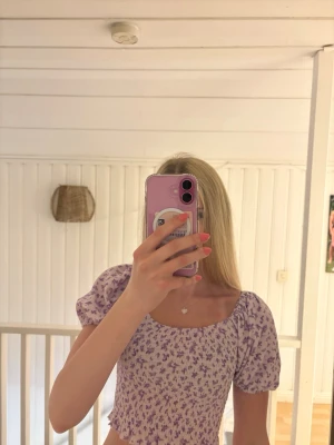 Lila blommig topp från Gina Tricot - Supersöt topp från Gina Tricot i ljuslila med blommigt mönster. Modellen har puffiga korta ärmar, croppad passform och smockad struktur som ger en snygg och bekväm look. Perfekt för dig som gillar trendiga och feminina plagg.