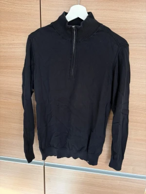 Svart halvzip tröja med hög krage - Stilren svart tröja med halv dragkedja och hög ribbad krage. Långärmad modell med ribbade muddar vid ärmslut och nederkant. Perfekt till jeans eller chinos för en clean look.
