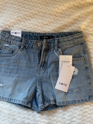  jeansshorts från LMTD - Säljer ett par ljusblå jeansshorts från LMTD med klassisk femficksmodell, bälteshällor och slitna detaljer framtill. Helt nytt, orginalpris 400