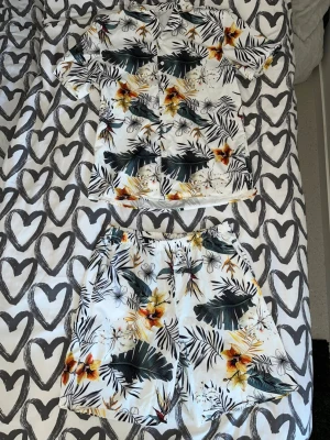 Blommigt set - Säljer ett snyggt tvådelat set med kortärmad skjorta och shorts i vitt tyg med tropiskt blommönster i grönt, gult och orange. Perfekt för sommaren! Skjortan har knappar framtill och krage. Materialet känns lätt och luftigt. Aldrig använt