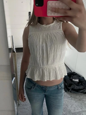 Vit ärmlös blus med volangkant - Söt vit blus i bomull med broderad spets upptill och volangkant nertill. Blusen är ärmlös och har en lätt rynkad struktur som ger en luftig känsla. Perfekt till jeans eller kjol för en fräsch look.