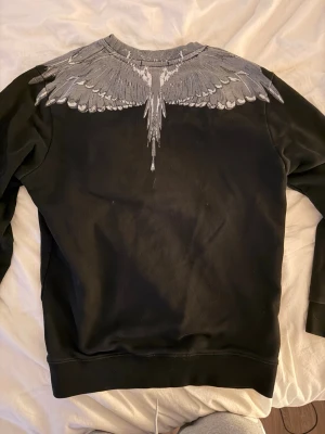 Svart sweatshirt från Marcelo Burlon - Säljer en svart sweatshirt från Marcelo Burlon med det ikoniska vingmönstret i grått och vitt runt halsen. Tröjan har rund halsringning, långa ärmar och ribbade muddar. Perfekt statement-plagg för dig som gillar streetwear och unika detaljer.