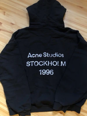 Svart hoodie Acne Studios Stockholm - Säljer en svart hoodie från Acne Studios med trycket 'Acne Studios STOCKHOLM 1996' i vitt på ryggen. Hoodien har klassisk huva och ribbade muddar. Perfekt för dig som gillar streetwear och vill ha något stilrent med en touch av Stockholm.