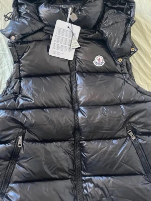 Svart dunväst från Moncler - Snygg svart dunväst från Moncler med glansig finish, två dragkedjefickor och Moncler-logga på bröstet. Västen har hög krage, avtagbar huva och stängs med dragkedja framtill. Perfekt för lager på lager och streetstyle.