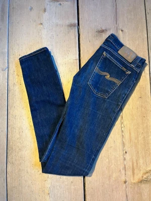 Mörkblå  jeans Nudie  w30 L34 - Snygga mörkblå skinny jeans från Nudie med klassiska kontrastsömmar och broderad bakficka. Jeansen har en smal passform och är tillverkade i mjuk denim med stretch för extra komfort. Perfekta för en trendig och avslappnad look.