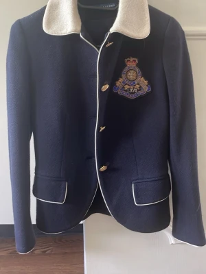 Marinblå kavaj från Ralph Lauren - Stilren marinblå kavaj från Ralph Lauren med vit kontrastkrage och vita passpoaler. Kavajen har guldfärgade knappar och ett broderat emblem på bröstet. Tillverkad i ett tjockare tyg som ger en exklusiv känsla. Perfekt för dig som gillar klassisk preppy-stil.