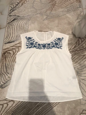 Vit blus med blå broderi - Vit ärmlös blus med vackert blått blommigt broderi över bröstet. Rund halsringning och lätt, luftig passform. Perfekt för varma dagar och enkel att matcha med jeans eller kjol. Använd endast ett fåtal gånger. Är öppen med prisförslag😊