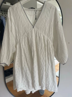 Vit broderad kort klänning - Söt vit kort klänning med v-ringning och broderade detaljer. Klänningen har trekvartsärmar med diskret spetskant och ett luftigt, löst fall. Perfekt för varma dagar och har fina detaljer längs halsringningen.