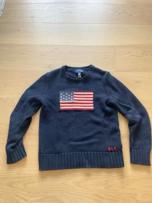 Mörkblå stickad tröja Polo Ralph Lauren - Mörkblå stickad tröja från Polo Ralph Lauren med amerikansk flagga broderad på bröstet och RL-broderi nertill. Tröjan har rund halsringning, långa ärmar och breda ribbade muddar. Perfekt för dig som gillar klassisk preppy stil.