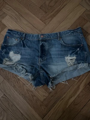 Lågmidjade korta jeansshorts - Säljer ett par blå jeansshorts med råa kanter och slitningar på framsidan. Shortsen har klassisk femficksdesign, knappstängning och är tillverkade i bomull. Perfekta för sommaren och jättesöta💕
