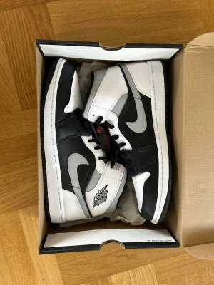 Nike Air Jordan 1 Mid svart/vit - Snygga Nike Air Jordan 1 Mid sneakers i svart och vitt läder med klassisk Swoosh på sidan och Air Jordan-logga vid ankeln. Skorna har svart yttersula och svarta skosnören. Perfekt för dig som gillar streetwear och ikonisk design.
