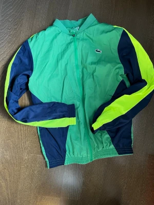 Grön vindjacka från Lacoste - Snygg grön vindjacka från Lacoste med dragkedja framtill och klassisk krokodil-logga på bröstet. Jackan har mörkblå och neongula detaljer längs ärmarna och elastiska muddar. Tillverkad i lätt och tunt material, perfekt för blåsiga dagar.