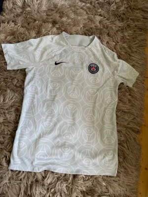 Vit PSG träningströja från Nike - Snygg vit träningströja från Nike med Paris Saint-Germain-logga på bröstet. Tröjan har ett diskret mönster i vitt och svart Nike-logga. Perfekt för fotbollsträning eller sportiga dagar.