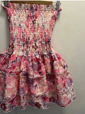 Rosa blommig kortklänning med volang - Supersöt kortklänning i rosa, lila och vit med blommigt mönster och volangkjol. Smockad överdel ger en tight passform upptill och kjolen är luftig och volangig. Perfekt för sommarens alla tillfällen!
