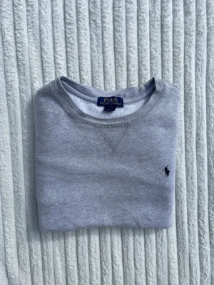 Grå sweatshirt från Polo Ralph Lauren - Klassisk grå sweatshirt från Polo Ralph Lauren med rund halsringning och diskret broderad logga på bröstet. Tröjan har lång ärm och är tillverkad i mjuk bomull för en skön och avslappnad look.
