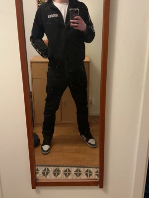 Svart hoodie från Hoodrich - Svart hoodie från Hoodrich med dragkedja och vit logga på bröstet. Klassisk modell med huva och fickor framtill. Perfekt för en avslappnad streetwear-look. Tillverkad i mjukt material för extra komfort.