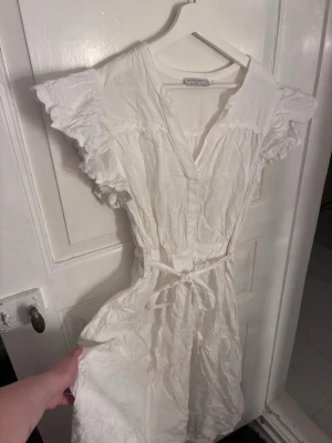 Vit klänning från Holly & Whyte - Söt vit klänning från Holly & Whyte med volangärmar, v-ringning och knytband i midjan. Tillverkad i luftig bomull, perfekt för varma dagar. Enkel och stilren design med romantiska detaljer.