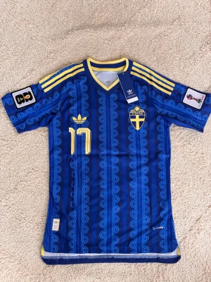 Blå Sverige fotbollströja Adidas - Retroinspirerad blå fotbollströja från Adidas med gula detaljer, Sveriges landslagsemblem och nummer 17 på bröstet. Tröjan har gula ränder på axlarna, V-ringning och officiella VM-märken på ärmarna. Tillverkad i lätt och ventilerande material.