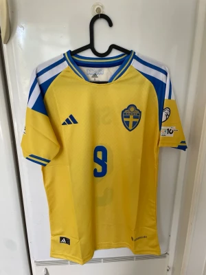 Svensk landslagströja Adidas gul/blå - Säljer en officiell svensk landslagströja från Adidas i gult och blått med nummer 9 på bröstet. Tröjan har korta ärmar, rund hals och broderad landslagssköld. Tillverkad i lätt och ventilerande funktionsmaterial, perfekt för fotboll eller träning.