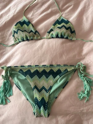 Turkos missoniliknande bikini - Turkos mönstrad missoniliknande bikini. Köpt på vinted och står ingen storlek men skulle säga s, kanske xs. Materialet är både vanligt baddräktsmaterial men mönstret är mer tyg/tråd material. Aldrig använd av mig själv, som sagt köpt på vinted men passar ej så jag säljer vidare. Underdelen är lågmidjad. Skriv gärna vid frågor eller prisförslag!