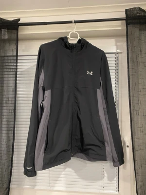 Svart och grå träningsjacka Under Armour - Snygg träningsjacka från Under Armour i svart och grått med dragkedja, huva och logga på bröstet. Jackan har elastiska muddar och sidofickor. Perfekt för löpning eller utomhusträning. Tillverkad i lätt och funktionellt material.