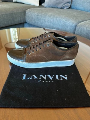 Lanvin cap toe DBB1 - Snygga bruna sneakers från Lanvin med ovandel i mocka och tåparti i mörkbrunt läder. Skorna har vita sulor och klassisk låg profil med snörning. Perfekta för dig som gillar stilrena och exklusiva sneakers. Uk 8 = passar 42-43