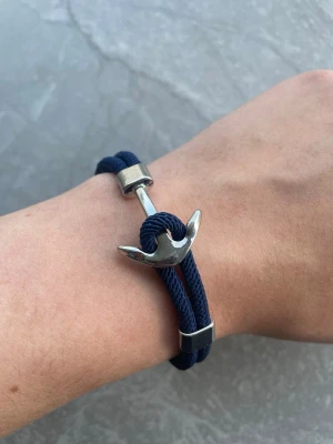 Marint armband med ankardetalj - Snyggt armband med marinblått rep och en silverfärgad ankare som lås. Armbandet har en stilren och nautisk känsla, perfekt för dig som gillar havsinspirerade accessoarer. Tillverkat i textil och metall för en cool och modern look.