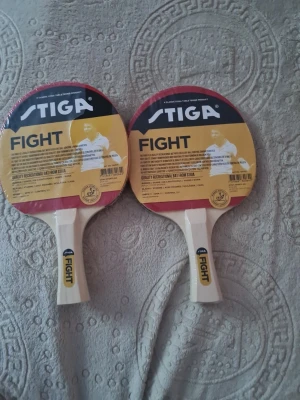 Stiga pingisracket och fodral - KOLLA ALLA BILDER. Två pingisracket av modellen Stiga Fight med trähandtag och röd/svart gummi, samt ett fodral i svart och orange med vit kant. Perfekt för dig som gillar bordtennis och vill ha bra utrustning från ett känt märke.