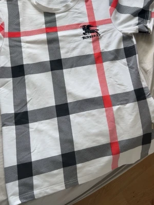 Rutig Burberry t-shirt i bomull - Snygg vit t-shirt från Burberry med klassiskt rutigt mönster i svart och rött. T-shirten har korta ärmar och rund halsringning. Tillverkad i mjuk bomull och har diskret Burberry-logga broderad på bröstet. Perfekt för en trendig och stilren look.