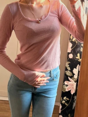 Ljusrosa ribbad långärmad topp - Söt ljusrosa ribbad topp med lång ärm och rundad halsringning. Toppen har en fin spetsdetalj vid halsen och är lätt croppad i modellen. Perfekt för dig som gillar en feminin och enkel stil.