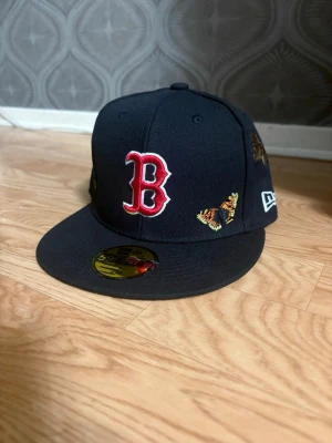 Svart Boston Red Sox keps New Era - Snygg svart keps från New Era med Boston Red Sox-logga i rött och vitt framtill. Kepsen har broderade fjärilar och bin på sidan för en unik look. Flat skärm och klassisk 59Fifty-modell. Perfekt för dig som vill sticka ut med din stil. Helt ny. Nypris kostar 550kr