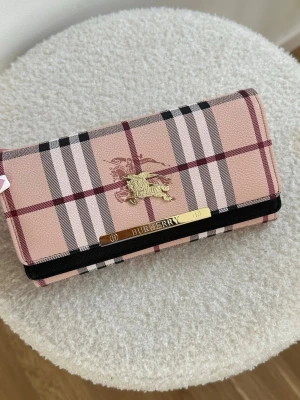 Rutig plånbok från Burberry - Säljer en helt ny snygg rosa/begie plånbok från Burberry med klassiskt rutmönster i svart, vitt och vinrött. Plånboken har guldfärgade detaljer, inklusive Burberry-loggan och en metallbricka framtill. Tillverkad i tåligt konstläder med elegant design och praktisk storlek.