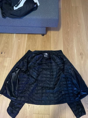 Svart pufferjacka från The North Face - Säljer en svart pufferjacka från The North Face med quiltad design och diskret logga på bröstet. Jackan har en lätt glansig yta och är tillverkad i syntetmaterial som håller värmen bra. Perfekt för kalla vinterdagar och riktigt snygg till streetwear-stilen.