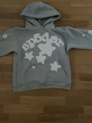 Grå hoodie Sp5der - Säljer en grå hoodie från Sp5der med stora vita stjärnor och logga framtill. Hoodien har magficka och huva, samt tryck på ena ärmen. Tillverkad i 100% bomull och har en avslappnad passform som passar perfekt till streetwear-stilen.