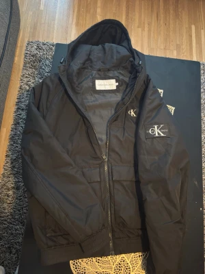 Svart vindjacka från Calvin Klein - Svart vindjacka från Calvin Klein Jeans med huva och dragkedja framtill. Jackan har två stora fickor, elastiska muddar och logotypdetaljer på bröstet och ärmen. Tillverkad i lätt, vindtätt material som passar perfekt för blåsiga dagar.