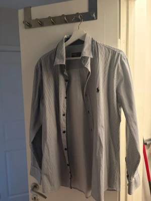 Randig skjorta från Polo Ralph Lauren - Säljer en klassisk vit skjorta med tunna svarta ränder från Polo Ralph Lauren. Skjortan har lång ärm, knappar framtill och den ikoniska loggan broderad på bröstet. Tillverkad i mjuk bomull och passar perfekt till både jeans och chinos.