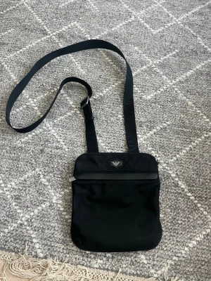 Svart axelväska från Emporio Armani - Snygg svart axelväska från Emporio Armani med justerbar rem och logotyp framtill. Väskan är tillverkad i nylon med detaljer i konstläder och har en stilren, rektangulär form. Perfekt för att bära det viktigaste med stil. Lite missfärgningar på metallen 