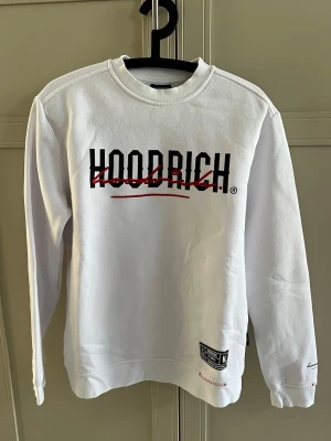 Vit sweatshirt från Hoodrich - Vit sweatshirt från Hoodrich med svart och rött tryck på både fram- och baksidan. Tröjan har rund halsringning, långa ärmar och ribbade muddar. Materialet är mjukt och bekvämt, perfekt för en avslappnad streetwear-look.