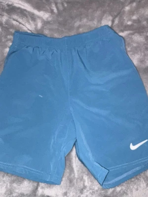 Blå Nike shorts  - Säljer ett par blå Nike shorts med elastisk midja och snörning på insidan för extra passform. Shortsen har en enkel design med den klassiska vita Nike-loggan på benet. Perfekta för träning eller chill dagar.