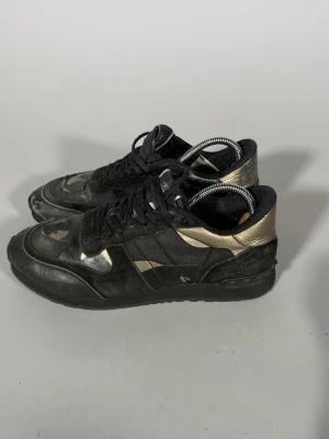 Svarta och guldiga sneakers - Snygga sneakers i svart med guldiga detaljer och klassiska ränder på sidan. Skorna har lågt skaft, snörning och en mix av mocka och textilmaterial. Perfekta för dig som vill ha en unik och cool stil. Slitna