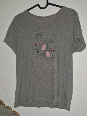 Tröja tshirt - Grå t-shirt i mjuk ekologisk bomull från. Framsidan har ett stilrent tryck med två abstrakta ansikten i svart, rosa och rött. Klassisk rund halsringning och korta ärmar. Perfekt för dig som gillar minimalistisk konst och sköna basplagg.