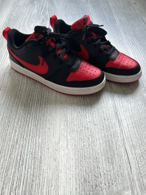 Nike svart-röda sneakers låga - Snygga låga sneakers från Nike i svart och rött med klassisk swoosh på sidan. Ovandelen är i läder och mesh, med svart snörning och vit sula. Perfekta för dig som vill ha en sportig och trendig look.