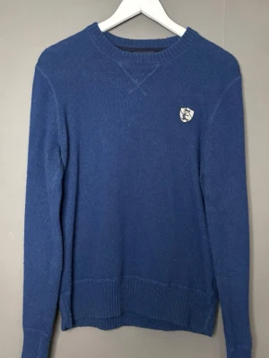 Blå stickad tröja från East West  - Mörkblå stickad tröja med rund hals och ribbade muddar. Tröjan har ett litet vitt emblem med blått tryck på bröstet. Klassisk och enkel design som passar till det mesta. Tröjan är i storlek M men sitter som S.