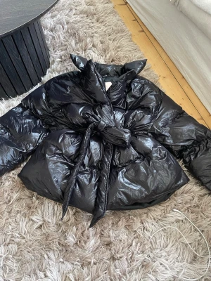 Svart pufferjacka med knytband - Säljer en svart pufferjacka med glansigt material och bred krage. Jackan har ett knytband i midjan som ger en snygg siluett och extra värme. Perfekt för kalla vinterdagar och riktigt trendig look.