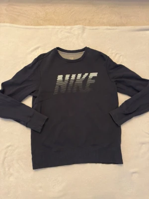 Svart Nike sweatshirt med tryck - Snygg svart sweatshirt från Nike med stort logotyp-tryck i vitt och grått på bröstet. Tröjan har rund halsringning, långa ärmar och ribbade muddar vid ärmslut och nederkant. Perfekt för en sportig och avslappnad stil.