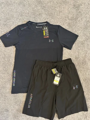 Svart träningsset från Under Armour - Snyggt svart träningsset från Under Armour med t-shirt och shorts. Båda plaggen har logga och textdetaljer i regnbågsfärger. T-shirten har rund hals och korta ärmar, shortsen har resår i midjan för skön passform. Perfekt för gymmet eller löprundan.