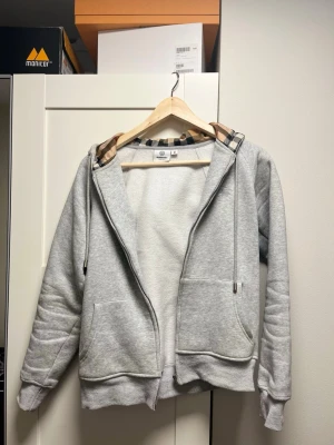 Grå hoodie med mönstrad huva - Snygg grå hoodie från Burberry med trasig kedja framtill och klassiskt rutmönster i huvan. Mjuk bomullsblandning, två fickor och ribbade muddar. Perfekt för en chill och stilren look. Eftersom dragkedjan är trasig kan den användas som en hoodie utan dragkedja